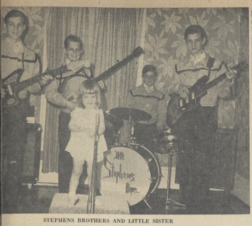 Four boys with musical instruments and their younger sister standing in front with a microphone.