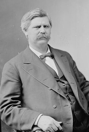 A black-and-white photograph of Zebulon B. Vance. He is sitting in a suit and bowtie and he has a large mustache.