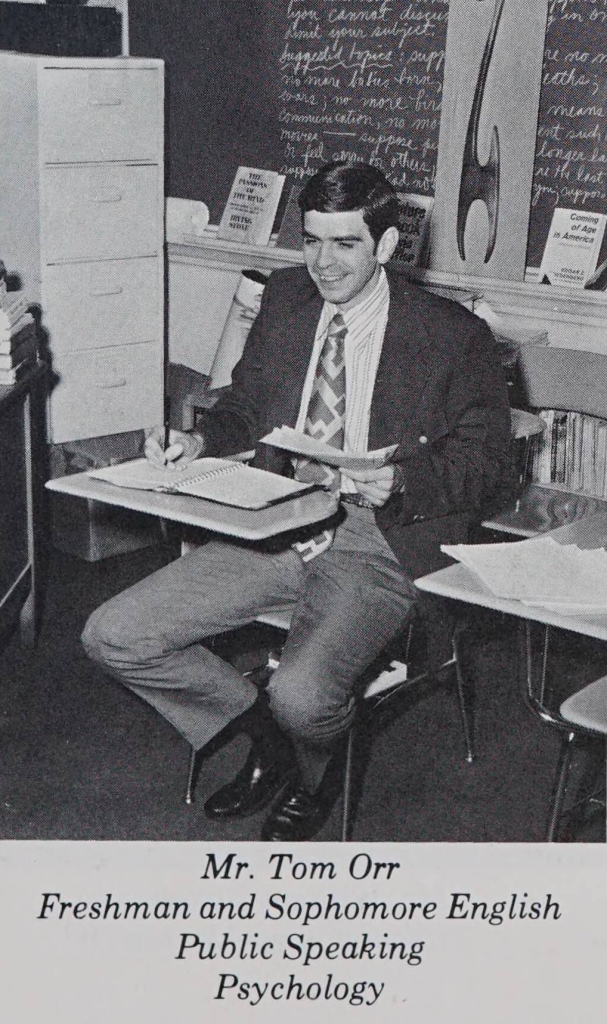 A young teacher sitting at a desk with a stack of papers in his hand. He is wearing a tie and suit jacket, and he seems to be laughing.