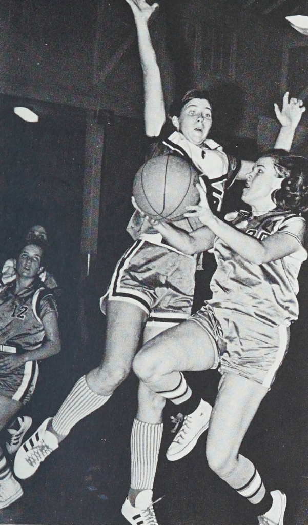 A black-and-white photo of two basketball players on the women's team jumping in the air. One is about to shoot the ball, and the other has her arms up to block.