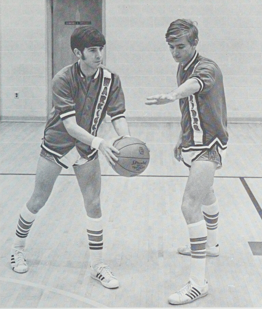 A black-and-white photo of two student basketball players posing with a basketball in high top socks and short shorts.