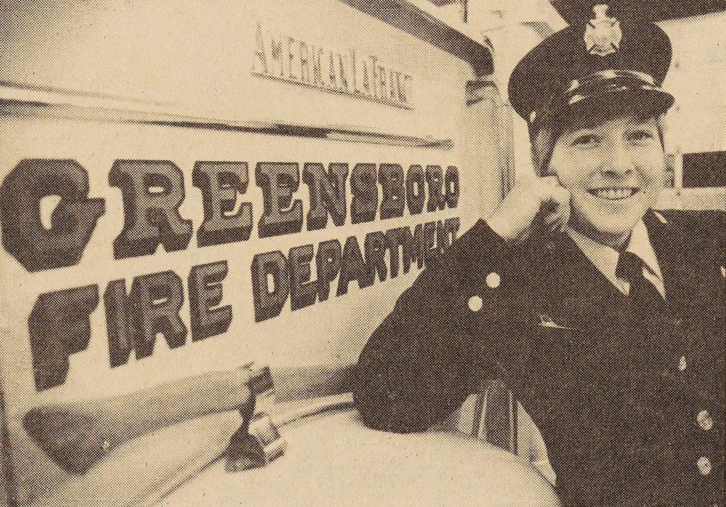 Image of a woman in her firefighter formal uniform posed next to a portion of a firetruck that reads "Greensboro Fire Department."