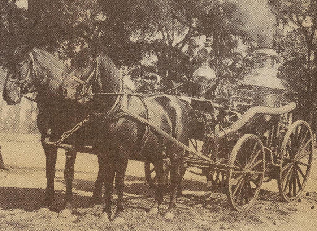 Two horses attached to a carriage carrying an old fire engine.