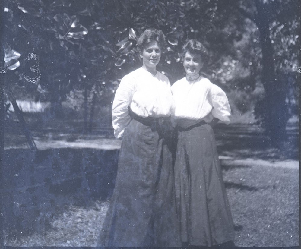 Two individuals dressed in light colored tops and dark colored skirts standing close to one another posing for the picture. Behind them are large magnolia trees.