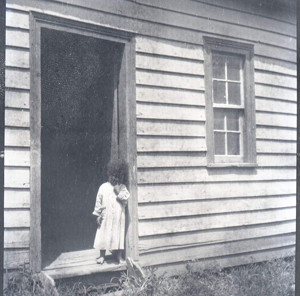 A small child standing in a doorway. The child is wearing a light colored dress.