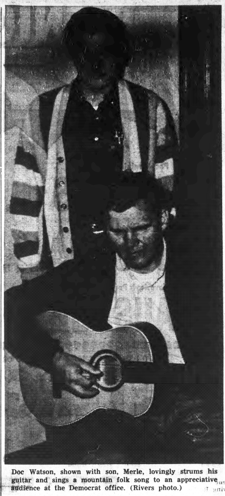 Photo of Doc Watson sitting down and playing guitar while his son stands behind him.