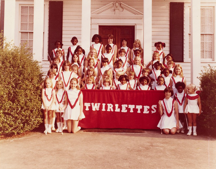A color portrait of a dance team posing in front of an old white house. The team consists of about 20 children wearing white uniforms and holding batons. The front row is holding a red banner that says "Twirlettes."