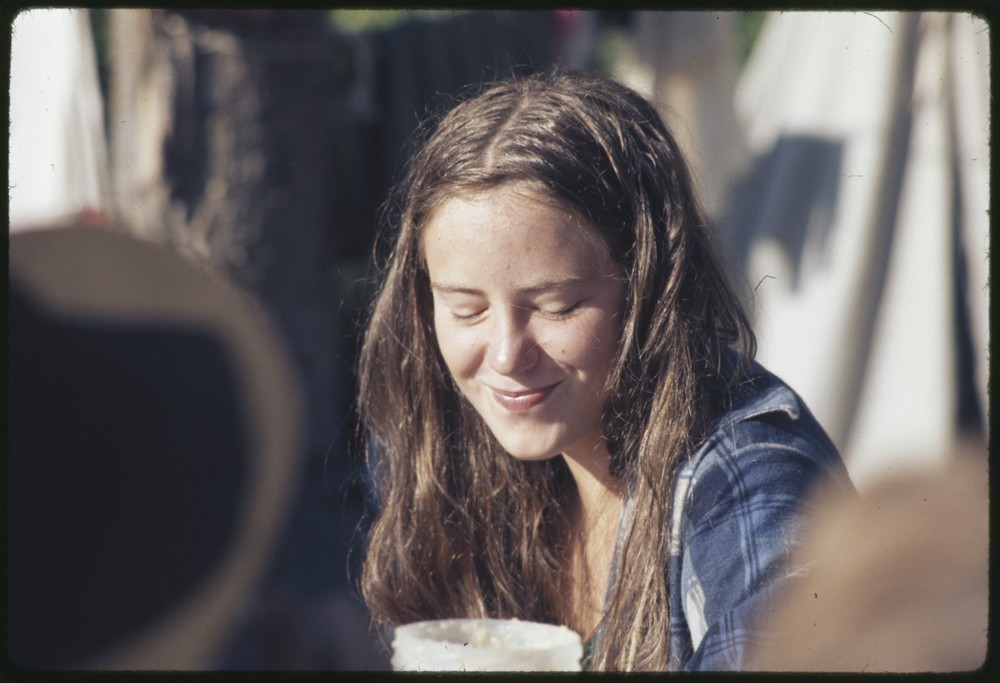 Younger individual with long brown hair sitting down and holding a cup.