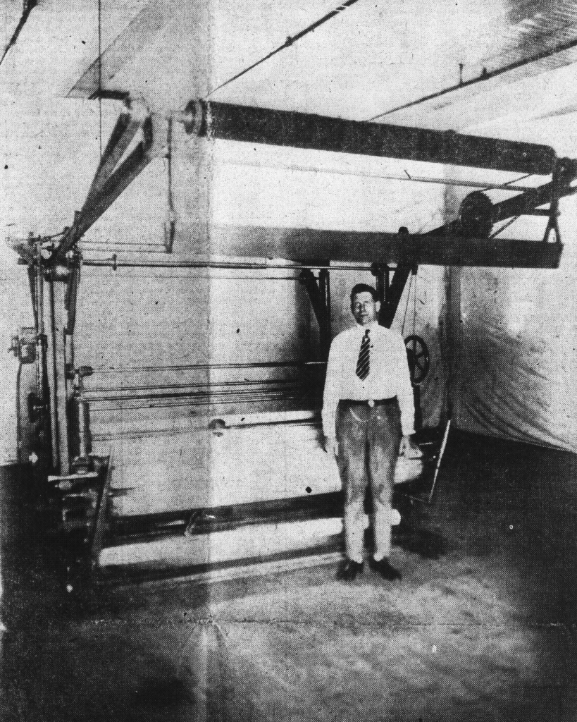 A black-and-white photo of an adult in a white button-down shirt, slacks, and tie standing in front of a large roller machine.