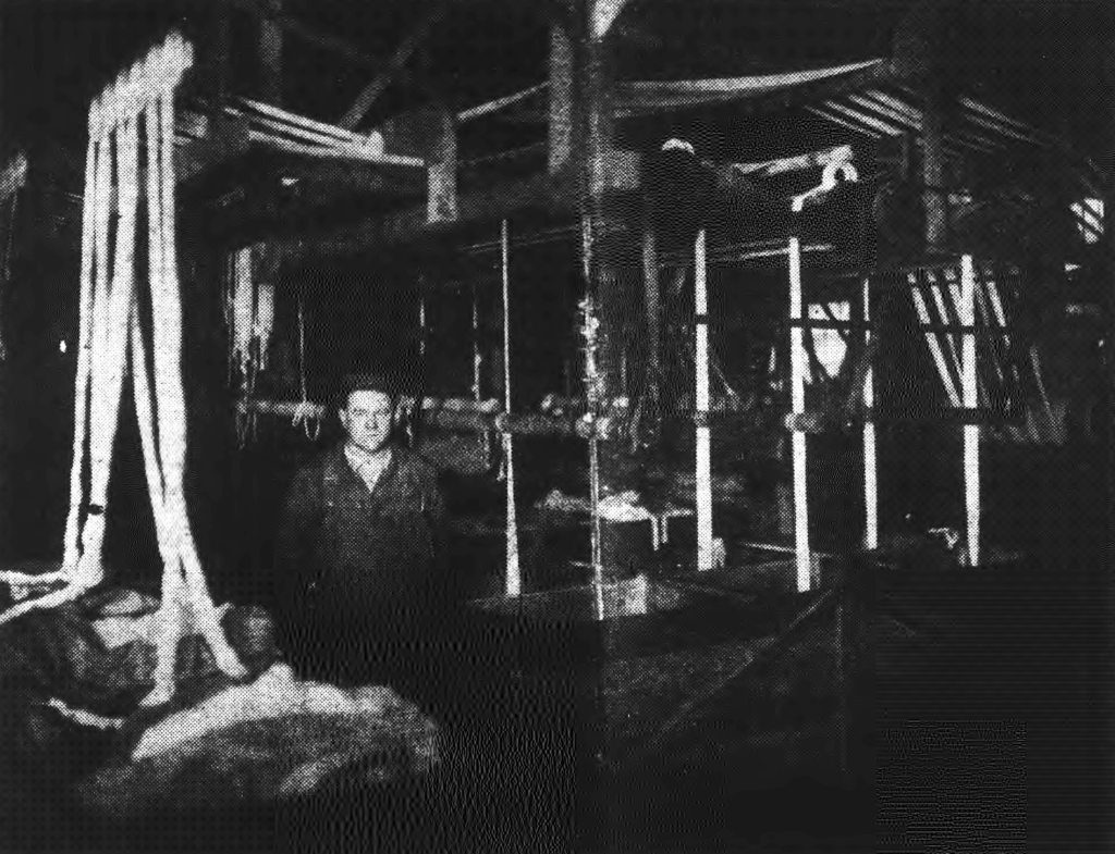 A black-and-white photo of an adult in a work shirt standing next to dye vats in a textile mill. The vats have long white fibers running out of them up to the ceiling.