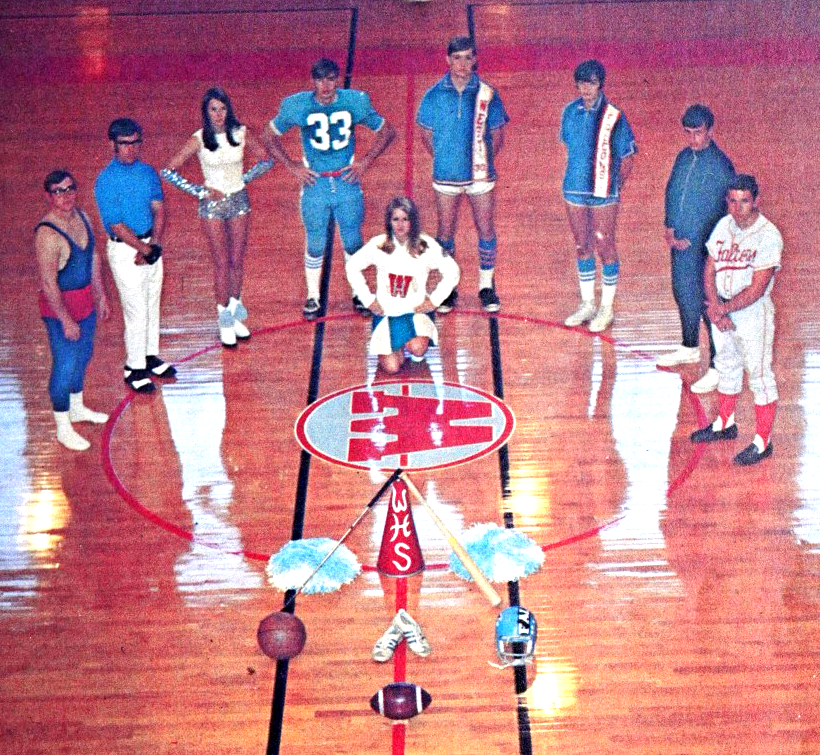 A color photo of representatives from several sports teams standing in a semi-circle around a cheerleader and some items in a school gym. The items include pom poms, a megaphone, a football, basketball, and baseball bat. Most of the players are wearing blue and white uniforms. 