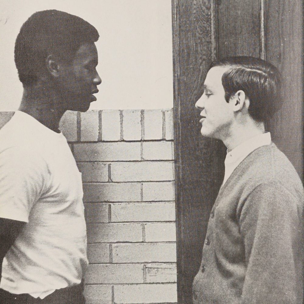 A black-and-white photo of a Black teenager on the left facing a white teenager on the right. Both are standing in profile against the hallway of a high school.