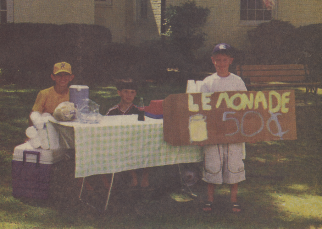 A color photo of two children sitting at a lemonade stand and a third child holding a cardboard sign that reads, "Lemonade, 50 cents."