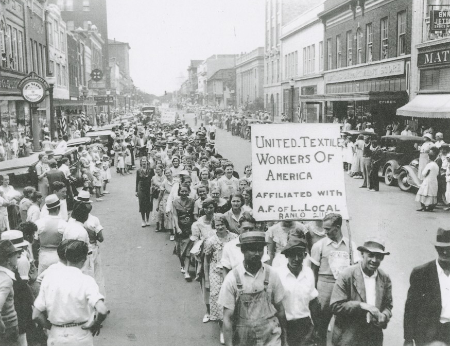 A black-and-white photo of textile workers marching down a public street. Near the front, a group of protestors holds a sign that reads, "United Textile Workers of America, Affiliated with A.F. of L. Local, RANLO 2118."
