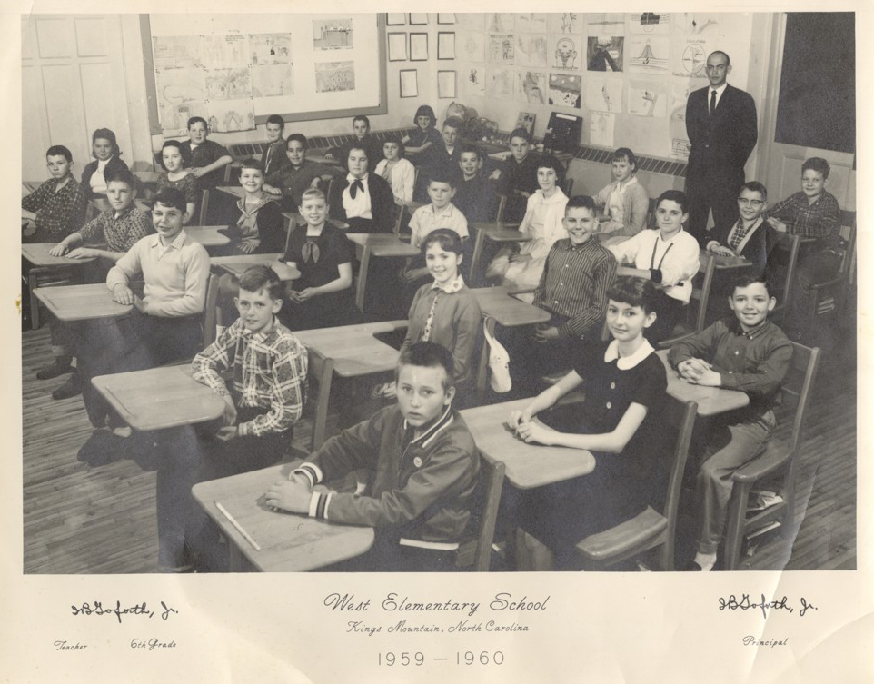 A classroom of white children sitting at desks and looking at the camera. Standing in the back of the room is their teacher/principal in a suit and tie.