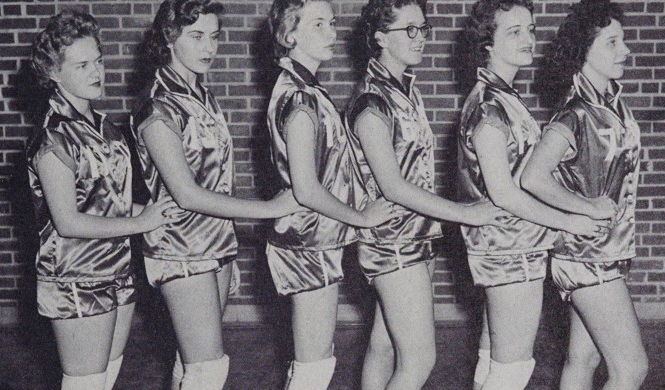 A black-and-white photo of six students wearing metallic dance uniforms and standing in a line, holding onto the hips of the person in front of them.