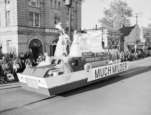 A parade float with young adults in dresses standing on top. On the side is a large sign that says 