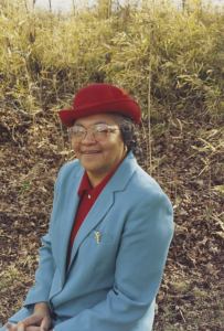 A portrait of an older woman in a blue blazer and red hat.