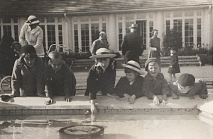 Six white children leaning over the edge of a grand fountain. In the background, three adults are talking, and one Black woman is standing with another white child.