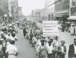 A black-and-white photograph of workers marching down a city street in Gastonia. One worker is holding up a large sign.