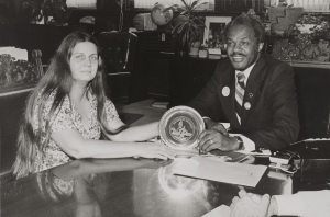 A white woman with long hair sitting across a table from a Black man in a suit. They are both holding onto a circular award between them on the table.