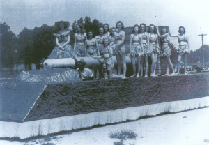 A blue-tinted photograph showing a parade float with about 12 young people riding it. They are dressed in tobacco-leaf skirts and bras.