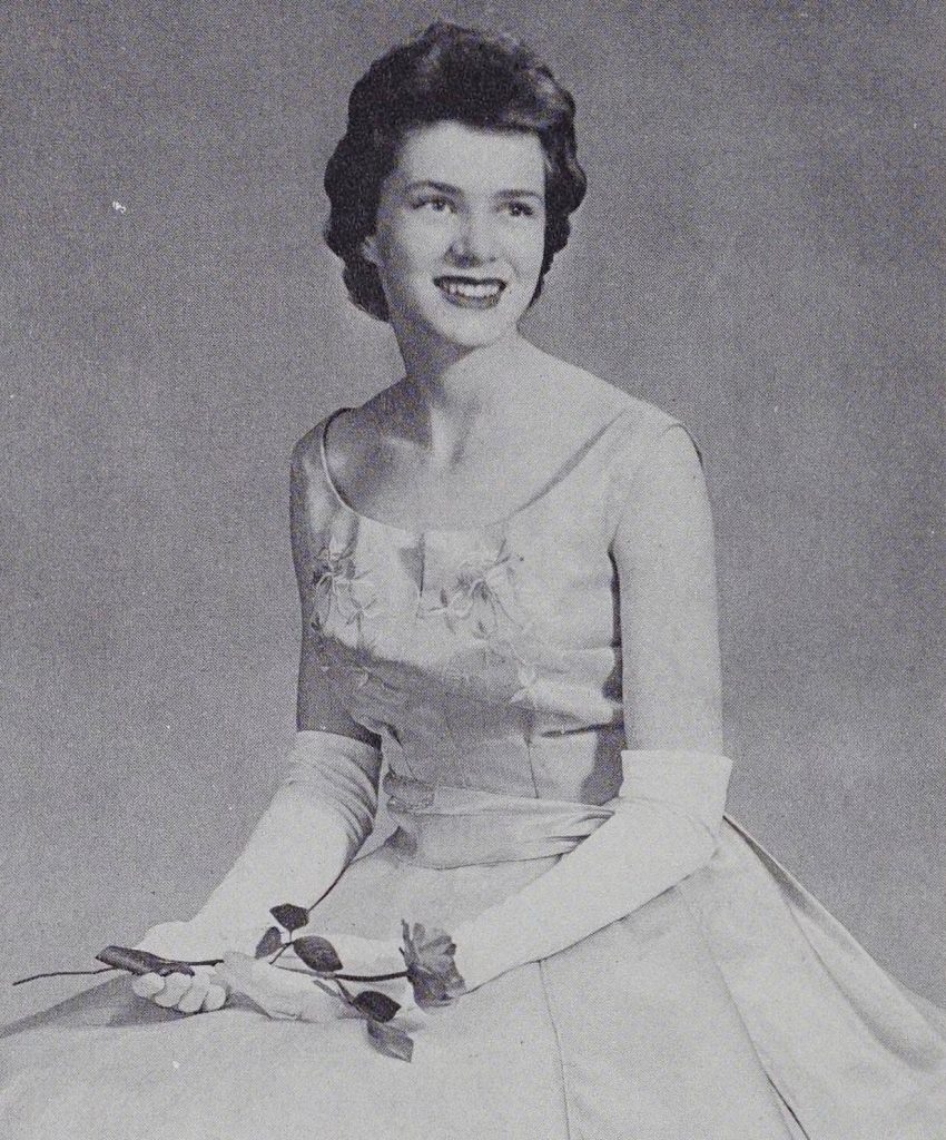 Individual in a light colored dress and long white finger gloves holding a rose while sitting in a chair.