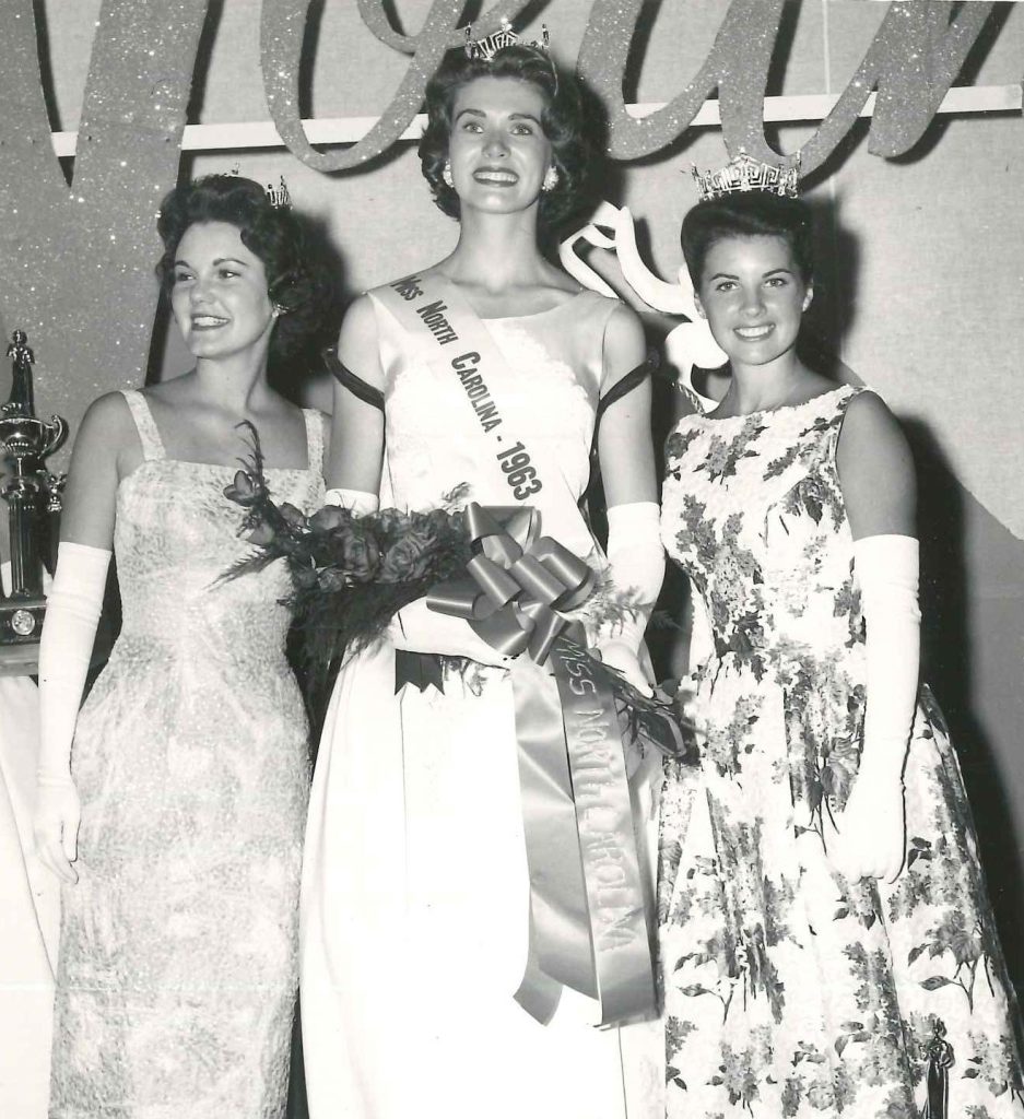 Three individuals standing together on a stage. Each person has on a long evening dress, long white finger gloves, and a crown. The person in the middle is holding a bouquet of roses and is wearing a sash that reads: Miss North Carolina - 1963.