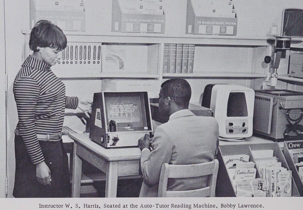 Individual sits at a desk with an Auto-Tutor Reading machine. Person stands to the left of the desk helping the person sitting to use the machine.