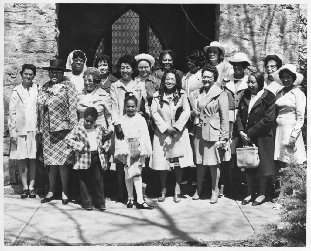 Black-and-white portrait of Episcopal Church Women and guests standing in front of the church wearing formal attire. 