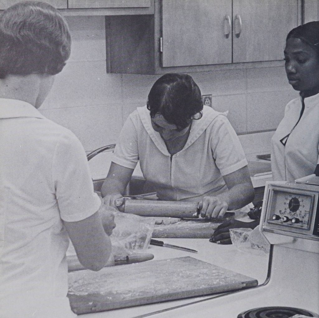 Two individuals looking at a third person using a rolling pin on cookie dough.