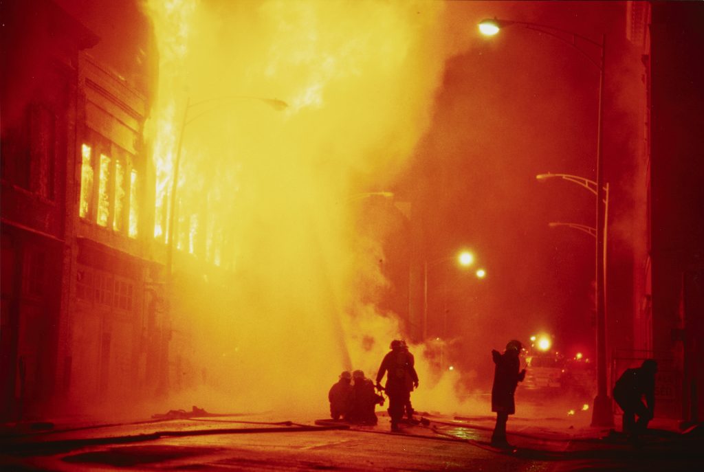 Two fire fighters hosing a building on fire. Two additional fire fighters standing near them. One fire fighter on the right side of the photograph pointing in the direction of the photographer. Unidentified individual bent over on the sidewalk near the pointing fire fighter.