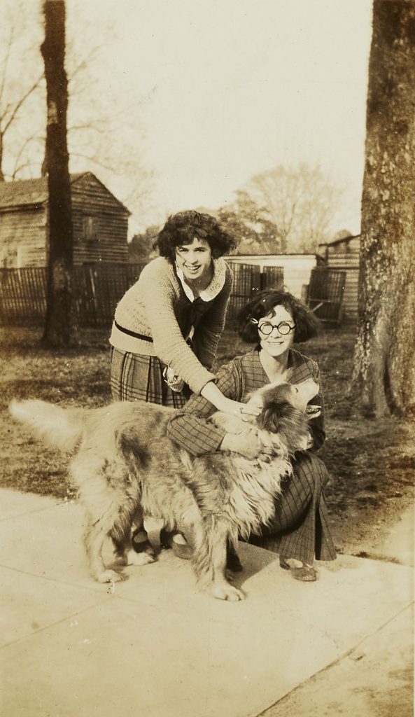 Two individuals petting an older golden retriever. One person is crouching and the other is bending over, both are looking at the camera and smiling.