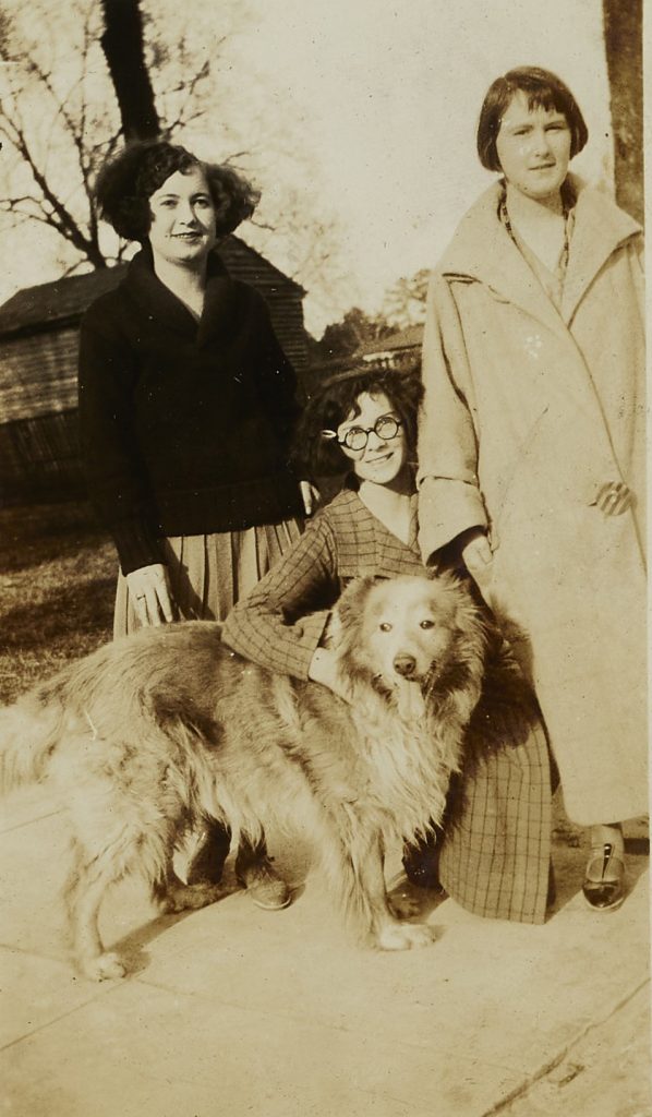 Three individuals and a dog. Two of the individuals are standing up, looking at the camera. The third is crouched down, petting the dog and smiling at the camera.