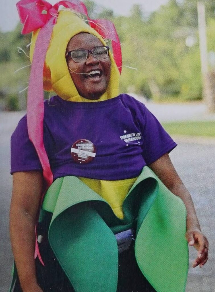 Student with glasses wears a corn costume with a pink bow on top. Over the corn costume is a purple t-shirt.