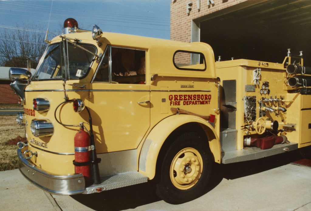 Side view of a yellow fire truck. The text to the right of the driver's door in red reads: "Greensboro Fire Department."