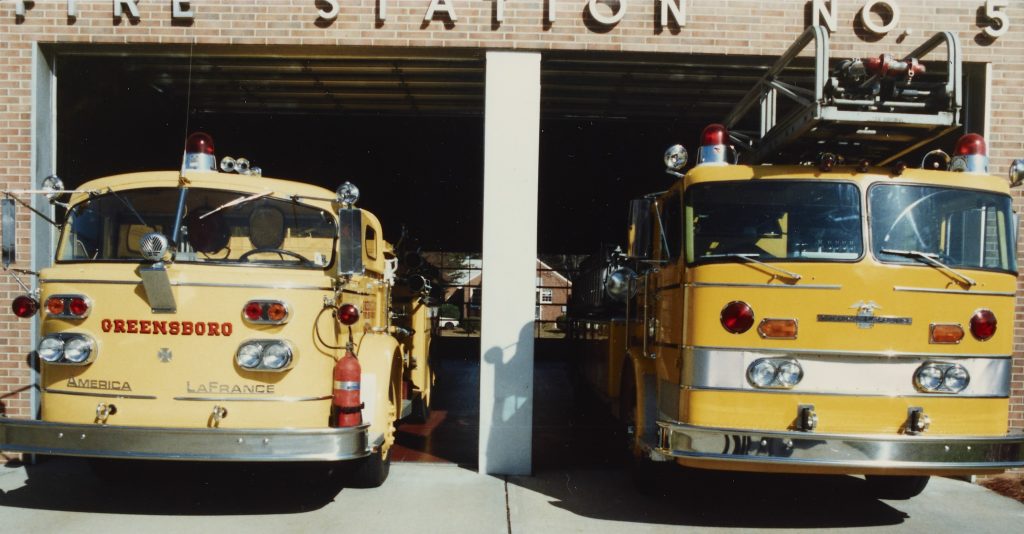 Front of two yellow fire trucks in their bays at Greensboro Fire Department's fire station number 5. The front of the left fire truck has "Greensboro" printed in red letters.