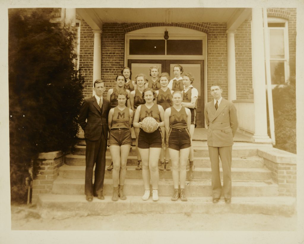 Twelve individuals standing on stairs in front of a brick building. The two outermost individuals on the bottom steps (on the left and right side) are dressed in suits while the rest are dressed in basketball uniforms. The uniforms consist of dark color shorts and a tank top with an "M" on it.
