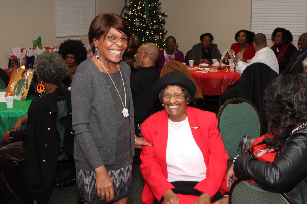 Two people smiling big. One person is standing on the left is dressed in a gray shirt and long sleeve cardigan. The second person, sitting, has on a black hat, white shirt, and red blazer.