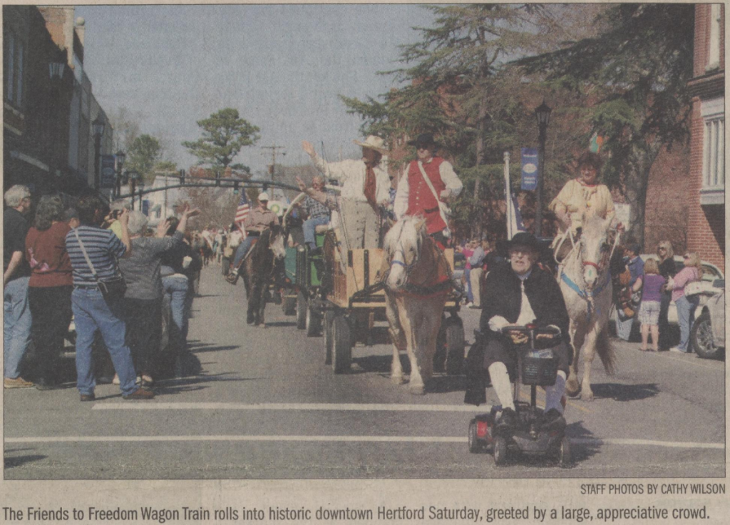 Photograph of individuals in a parade formation. At the very front of the parade is an individual riding a motorized scooter in a Quaker outfit. Behind that person some people are in buggies and wagons being pulled by horses and others riding on horses. Some people are waiving American flags. People are on the sidewalks taking pictures with their phones or waving to the people in the parade.

Below the photograph the caption reads: The Friends to Freedom Wagon Train rolls into historic downtown Hertford Saturday, greeted by a large, appreciative crowd.