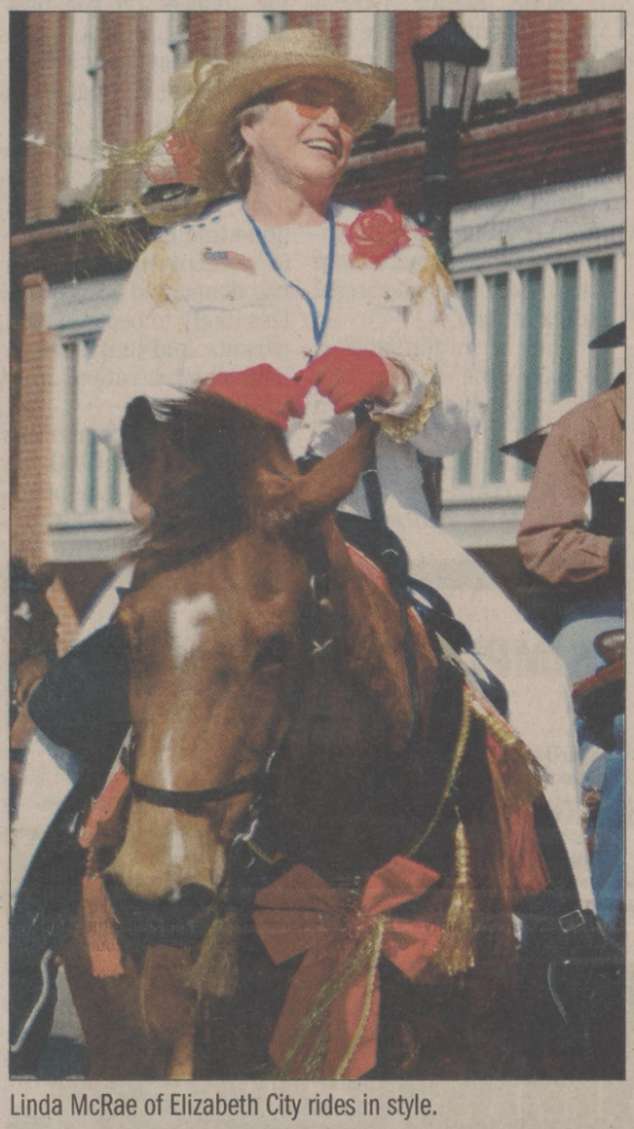 Individual wearing a wicker hat, white outfit with a red rose on their left shoulder, and American flag on the right side of their chest, and red gloves, riding a horse.

Below the photo the caption reads: Linda McRae of Elizabeth City rides in style.