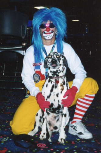 A person wearing a blue wig and yellow pants kneels on one knee holding a black and white spotted Dalmatian dog. 