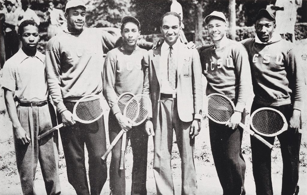 Individuals primarily in sportswear holding what appears to be tennis rackets posing and smiling for the camera.