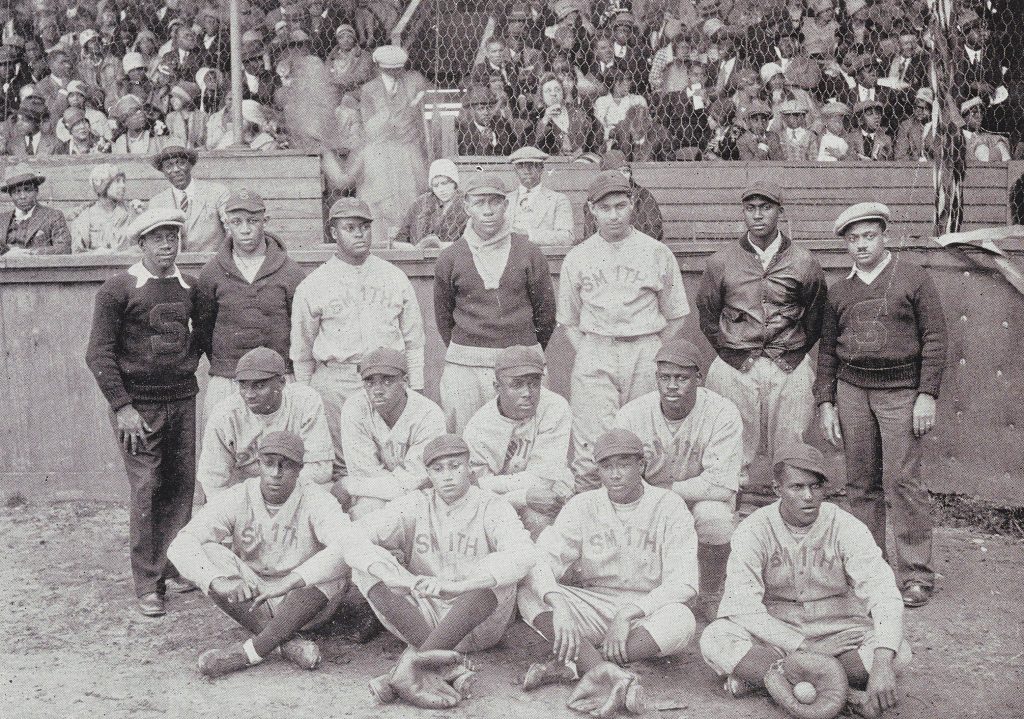 Photograph of the JCSU baseball team. In the background audience members sit on bleachers. In the forefront are team members in their uniforms and some with their gloves. Behind the players are individuals in sweaters and newsboy hats.