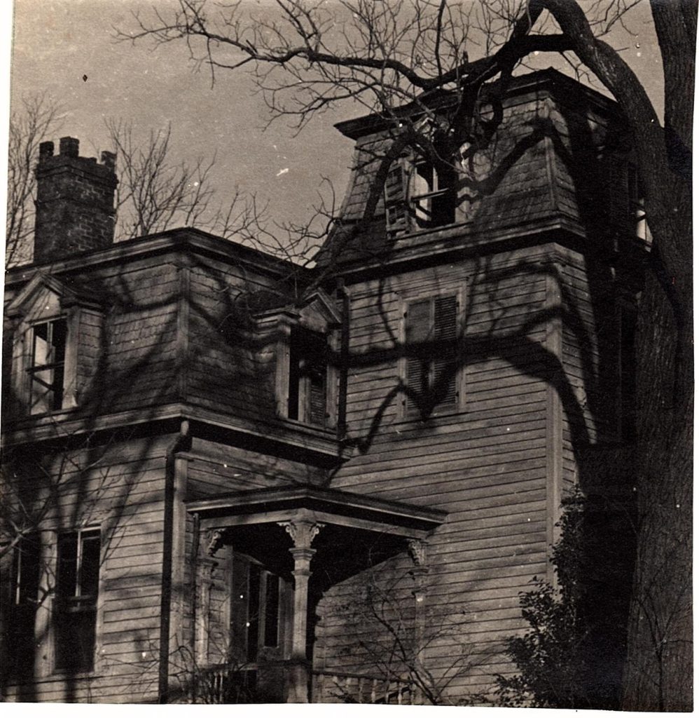 A black-and-white photo of a house built in a Victorian style. A tree with gnarled branches is in the foreground, casting a shadow on the manor.