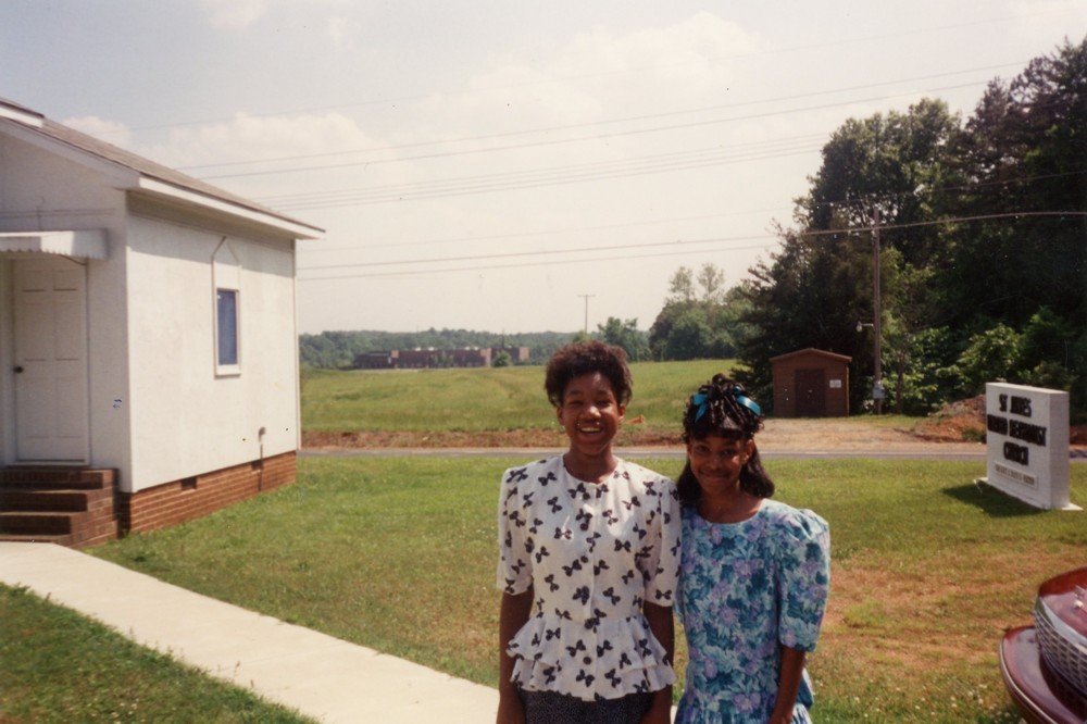 Two individuals dressed up standing next to one another. Behind them is a green field and brick building off in the distance. To their back left is a white building with brick base. To their back right is a sign with black letters that in-part reads: St. James [?] [?] Church.
