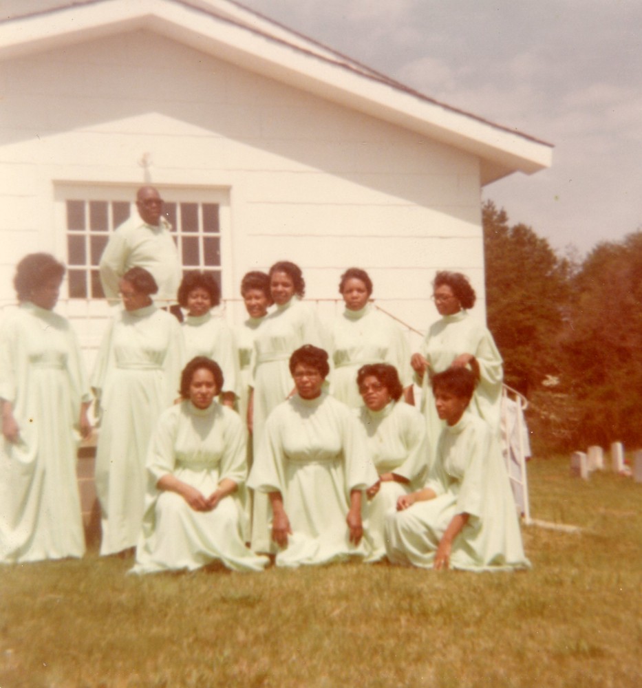 Individuals dressed in mint green dresses (and one individual in the very back with a mint green polo) pose for a photograph in front of a white building.