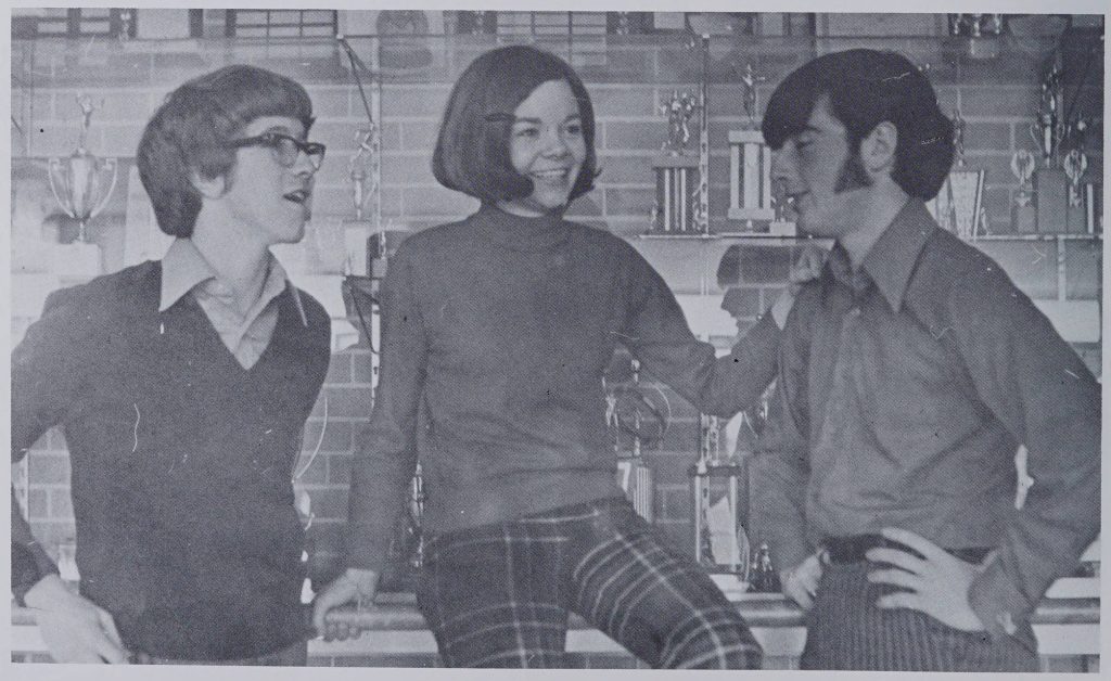 Three students in front of a trophy case. The individual in the middle is wearing a turtle neck shirt with pants and is smiling. The individuals on the sides are looking towards the person in the middle, both wearing long sleeved shirts with their hands on their hips.
