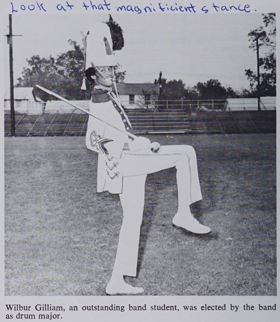 Individual dressed in a drum major uniform complete with a tall shako (hat) with tassle. The individual has one knee up high and a baton in their hand. 
