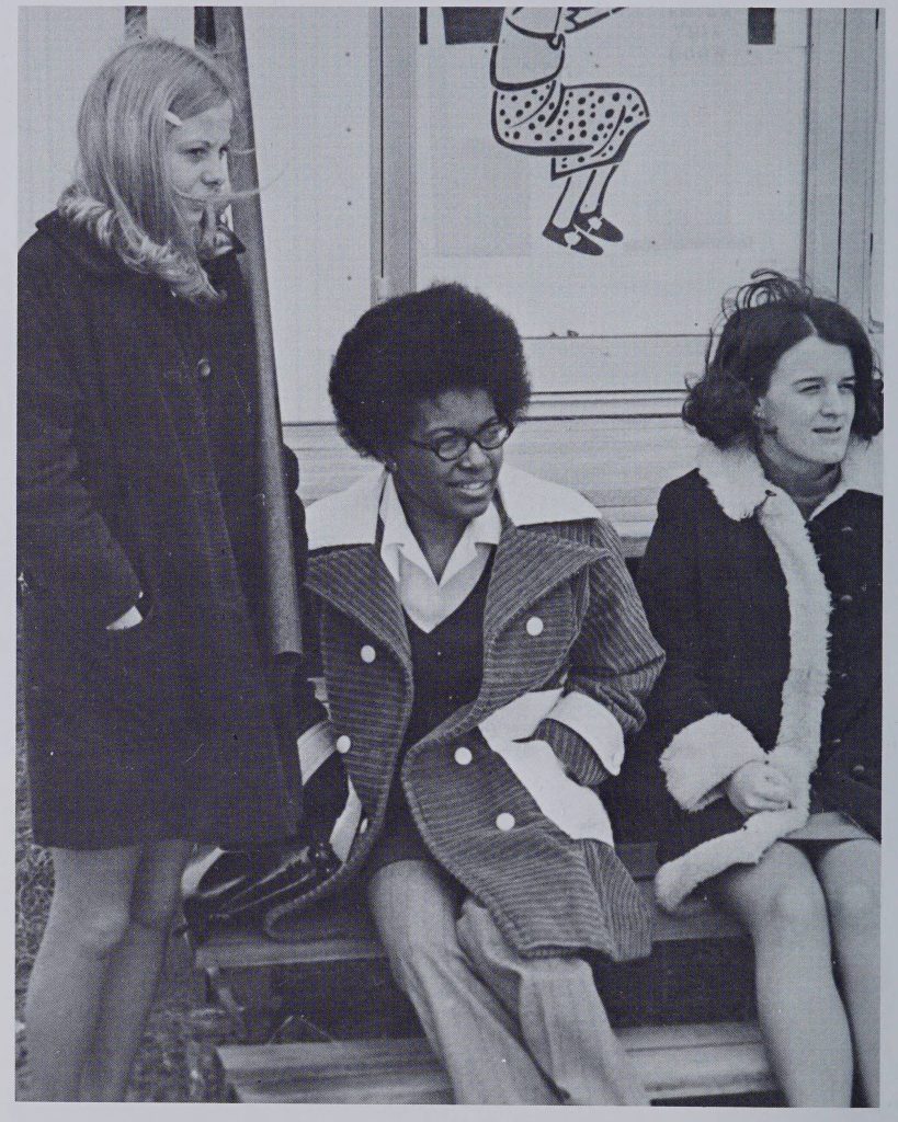 Three individuals photographed. The person on the left is standing while the other two are setting on some steps. The individual in the middle has on pants and a heavy coat.